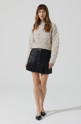 A woman is wearing a chunky, cream-colored cable knit sweater with long sleeves and a high neckline. She pairs it with a black leather mini skirt that features vertical pleats or panels. She completes the outfit with black pointed-toe shoes that have decorative embellishments on the front.